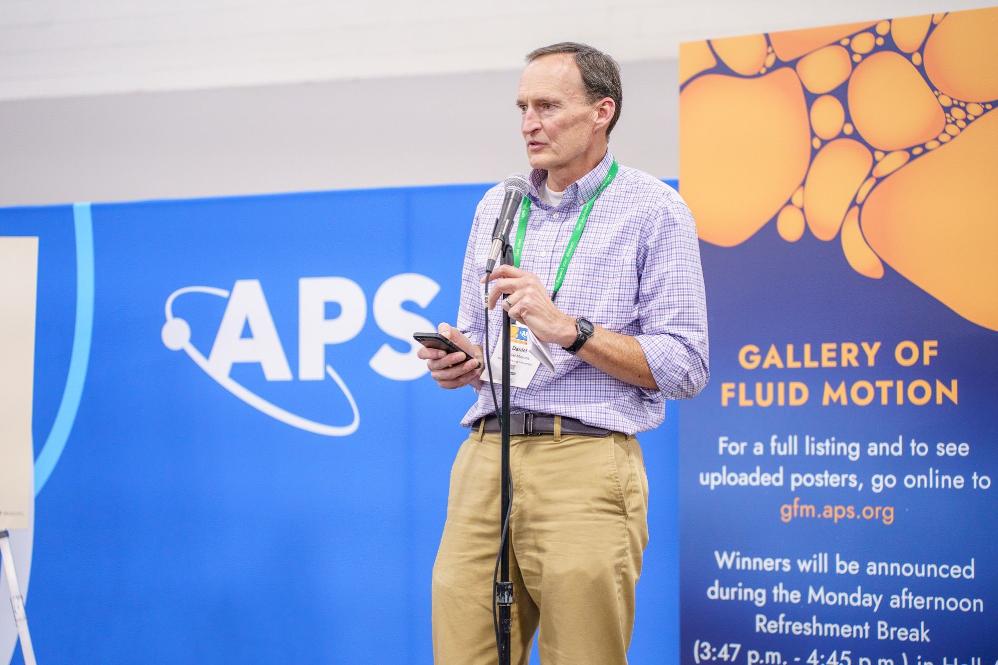 A man standing at a microphone in front of graphics for APD and Gallery of Fluid Motion